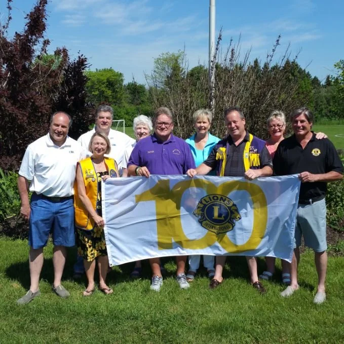 	
Mayor Steve Pellegrini standing outdoors on grass, holding a Lions Clubs banner with the number 100, celebrating an anniversary.
