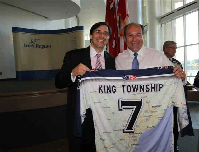 Two men in business attire holding a King Township hockey jersey with the number 7, inside a building with a York Region sign behind them.