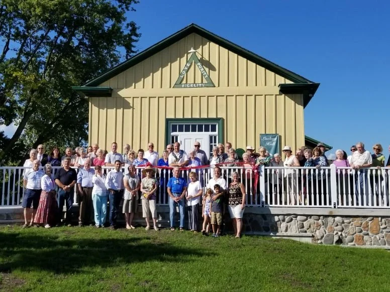A large group of people gathered with Mayor Steve Pellegrini in front of a yellow building with a white porch, some holding a red ribbon. The building has a sign with crossed tools and the words 'FELICIT'. 