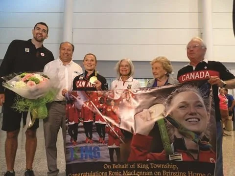Group of six people celebrating, holding a banner featuring a hockey player and a photograph of a woman, with some holding Canadian flags and flowers, indoors at an event.