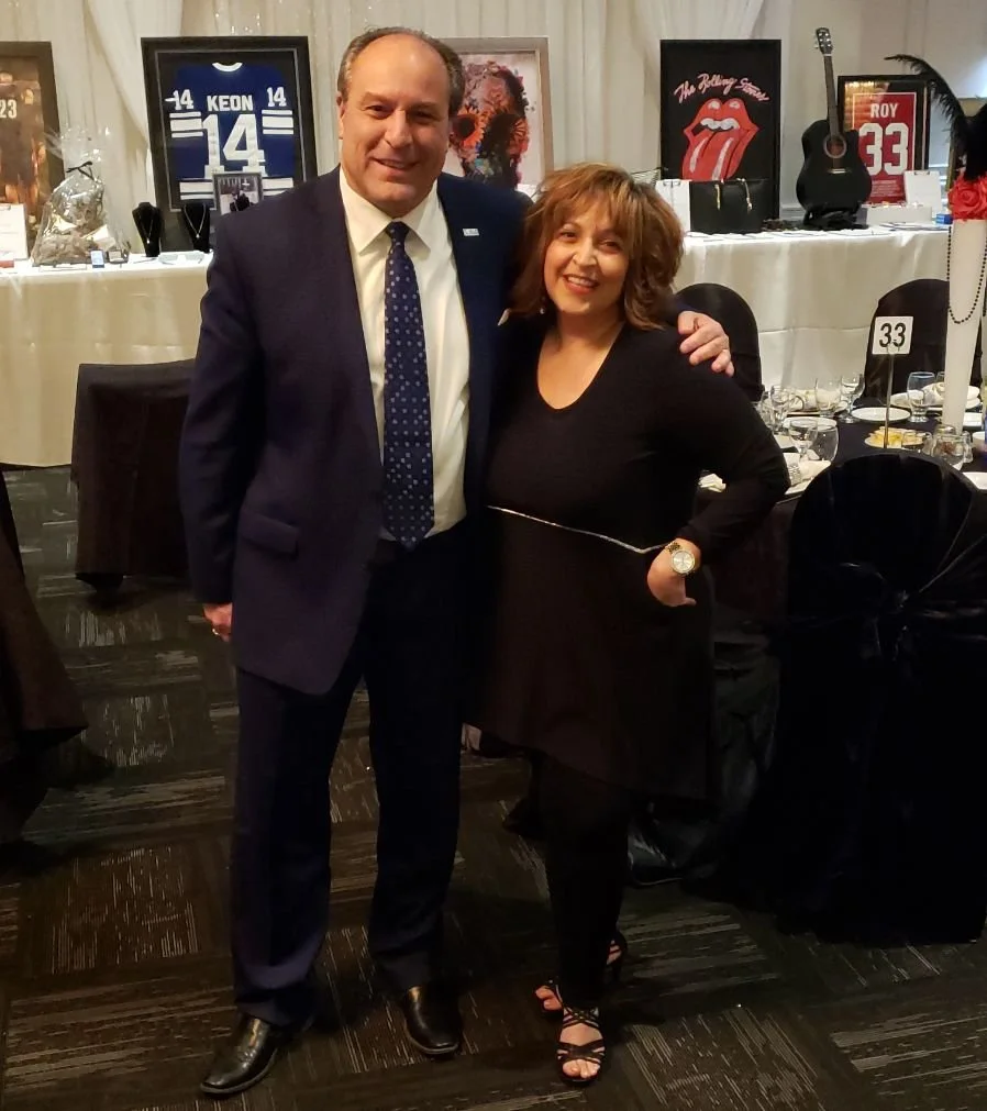 A man and woman posing together at an event, with tables decorated with memorabilia and artwork in the background.