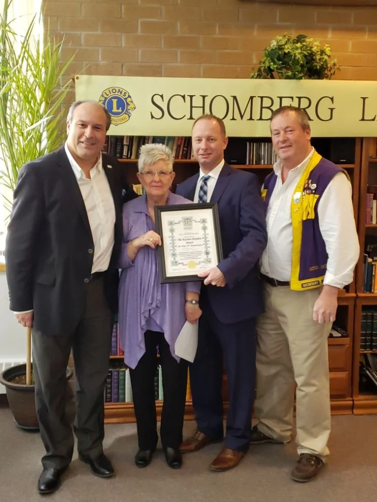 Four people standing together celebrating, one woman and three men, holding a framed certificate, in front of a bookcase with a banner that reads 'Schomberg Lions Club' and a Lions Club logo.