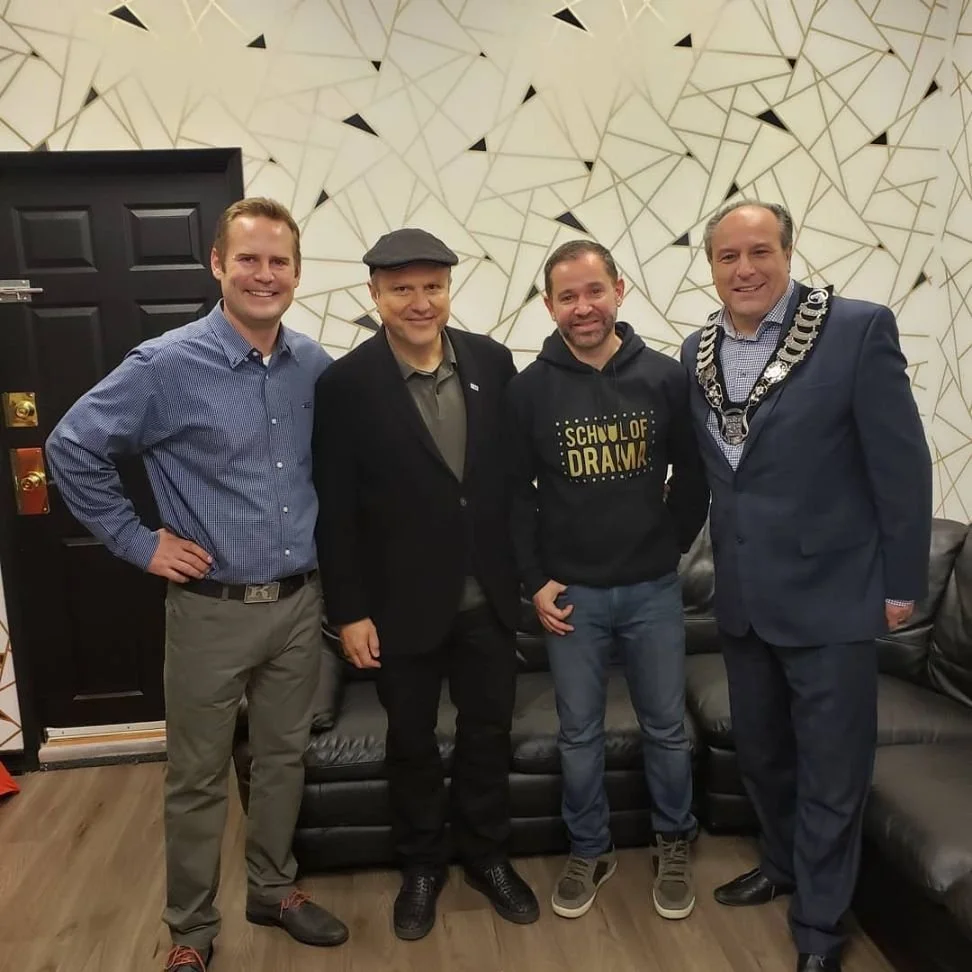 Four men standing in a room with a modern geometric wall design, a black door, and a black leather couch. One man wears a suit with a chain of office, indicating a formal or official event.