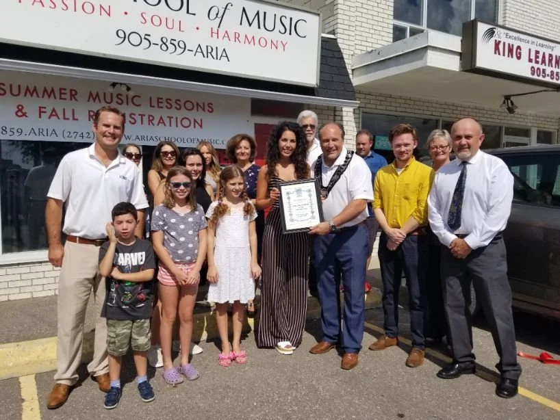 Group of people standing outside a music school, some holding a framed certificate, in front of a sign for the school.