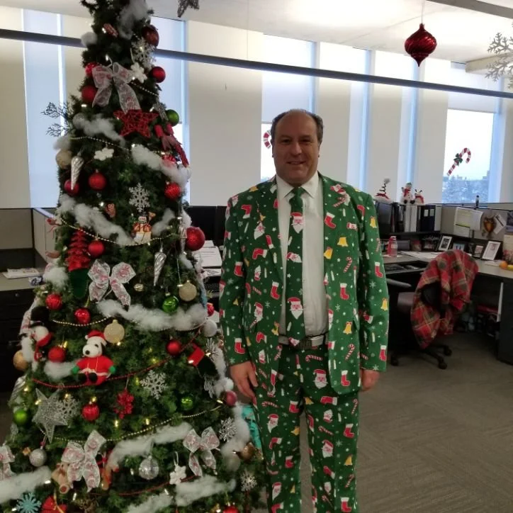 Mayor Steve Pellegrini in a Christmas-themed suit standing next to a decorated Christmas tree in an office setting.