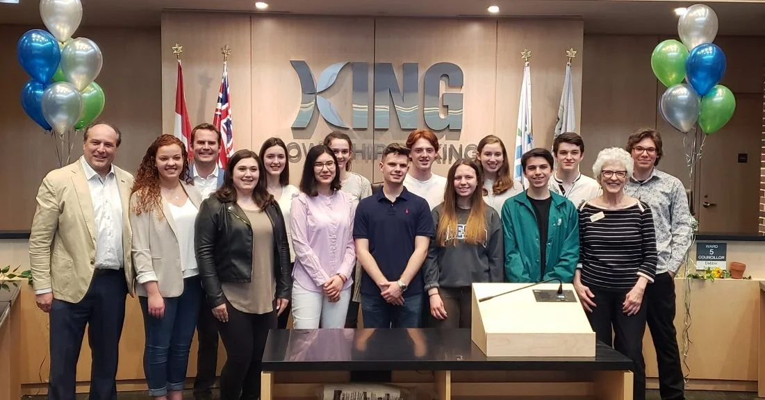 Group of young people and adults gathered in a room with a theater-style setup, standing in front of a wall with the large logo of XING, a professional networking company. The group is smiling, and there are colorful balloons on each side of the grou