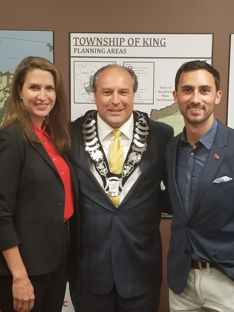Three people standing together in front of a township planning sign. The man in the center is wearing a chain of office, indicating he is a local official, with a woman on his left and a man on his right, all smiling.
