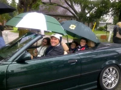 Four people sitting in a convertible car holding umbrellas during rainy weather. They are smiling and appear to be enjoying the outing.