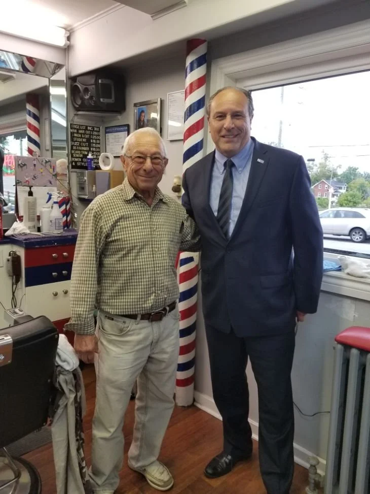 Two men standing inside a barber shop with barber poles in the background. One older man with glasses and a checkered shirt, and a younger man in a suit.