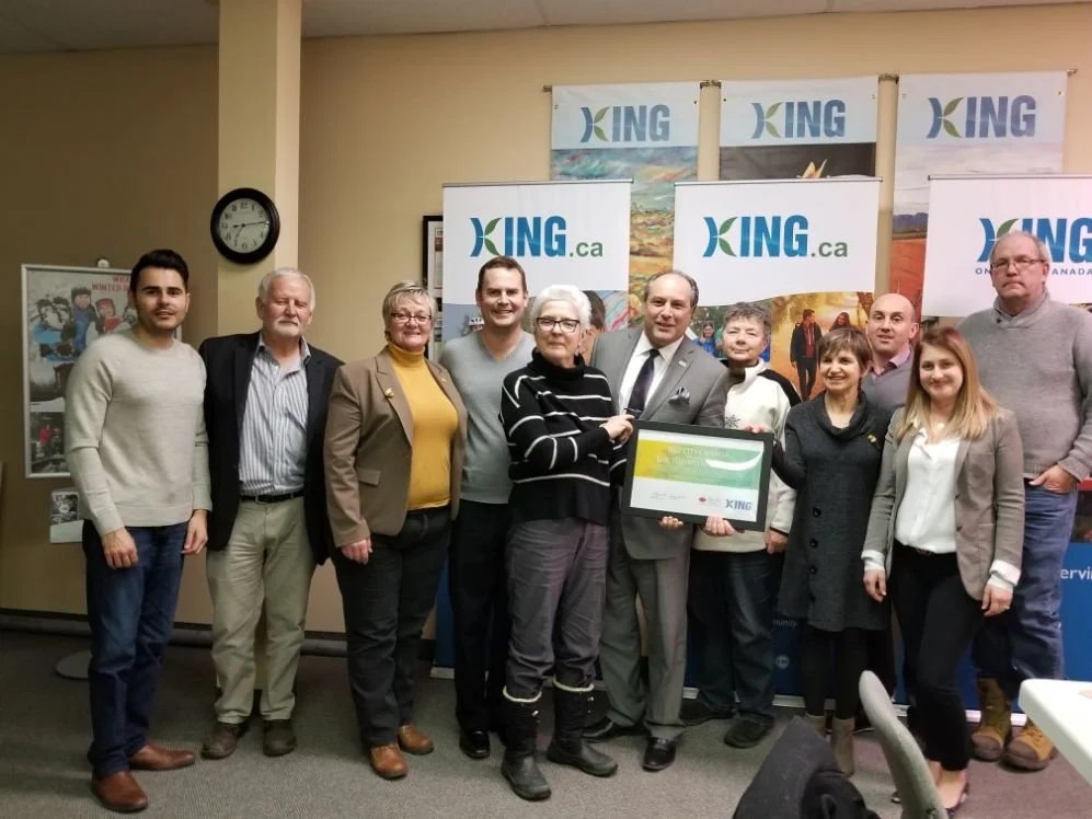 A group of eleven people standing in a room, holding a framed certificate or award, with banners behind them displaying the logo and website 'KING.ca'.