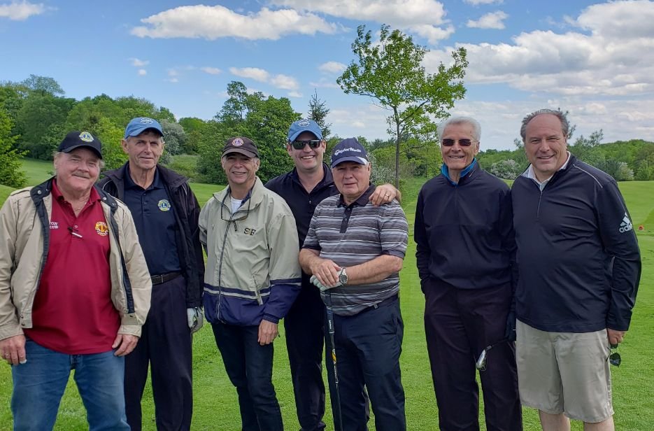 Seven men standing together on a golf course, smiling, with trees and a partly cloudy sky in the background.