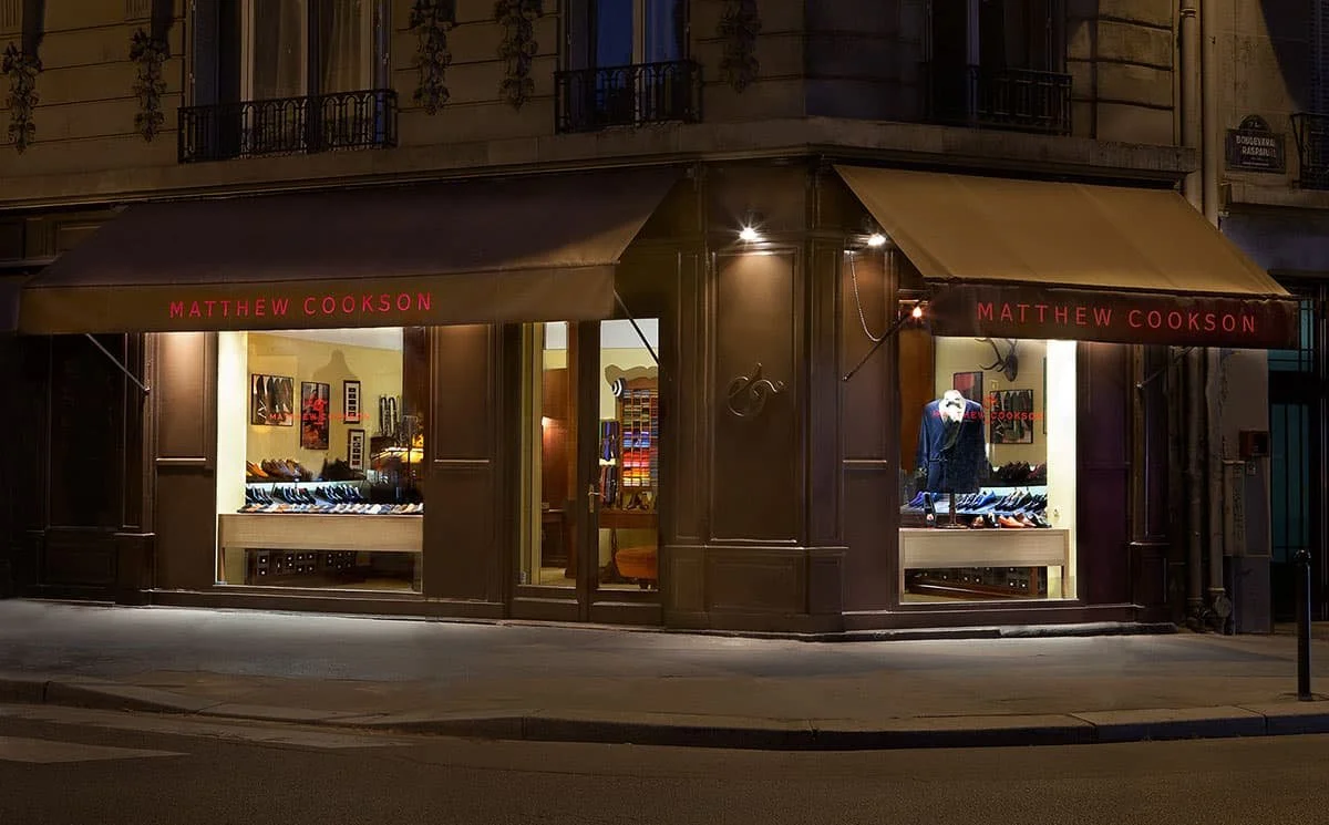 Nighttime view of a men's clothing store named Matthew Cookson with illuminated display windows showcasing shoes and clothing, located on a corner of a city street.