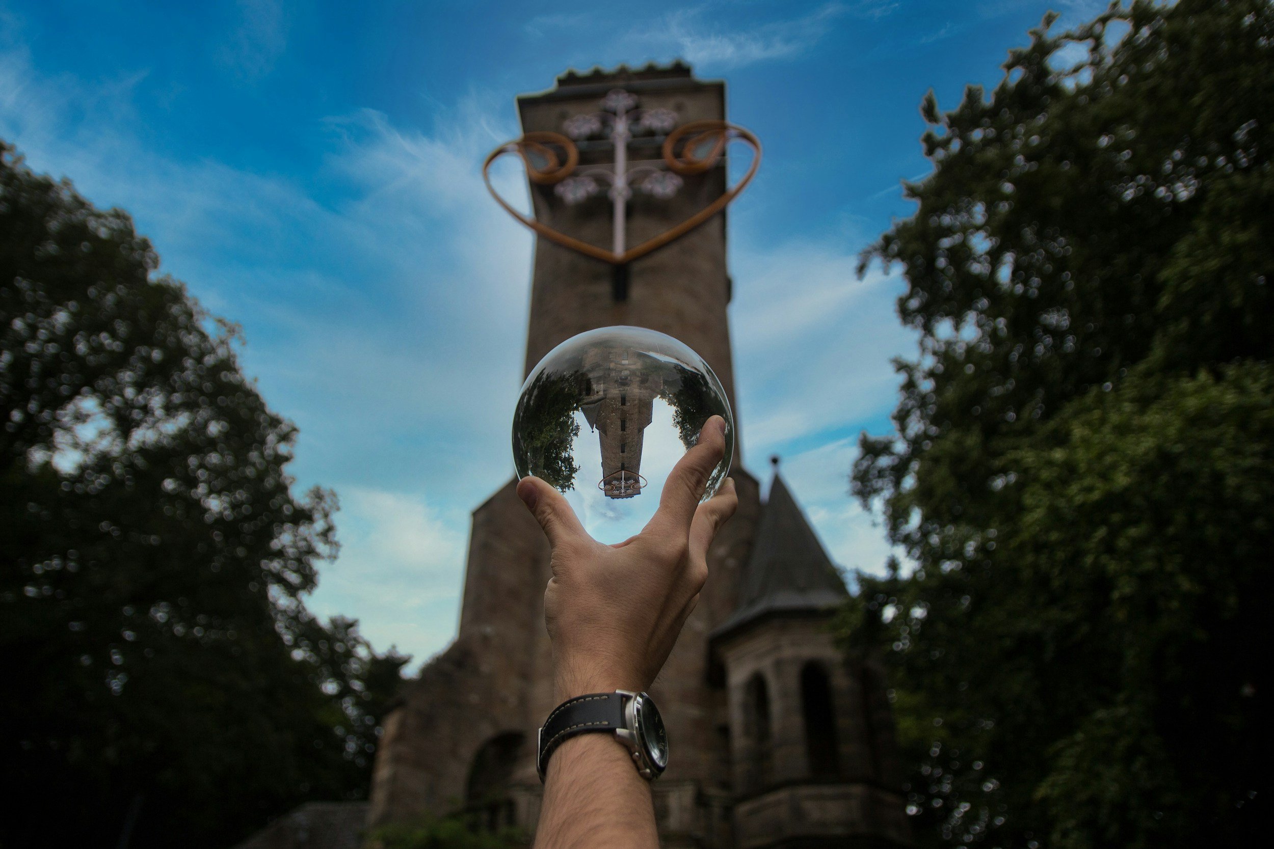 A person's hand holding a glass sphere in front of a tall stone tower with an iron cross at the top, framed by trees and a partly cloudy sky.