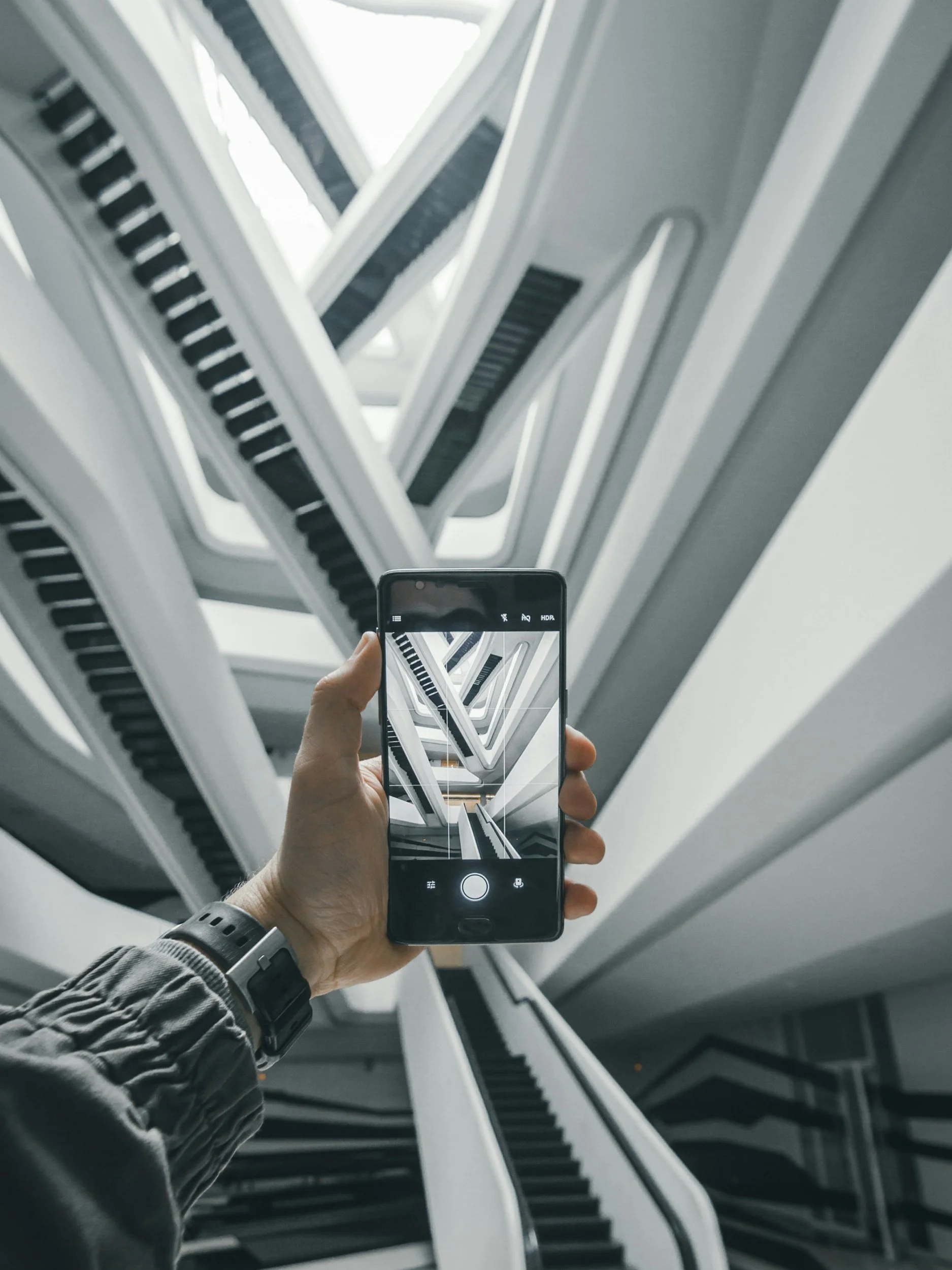 A person holding a smartphone capturing a photo of an upward view of a staircase with white railings and black steps, creating a repetitive pattern.