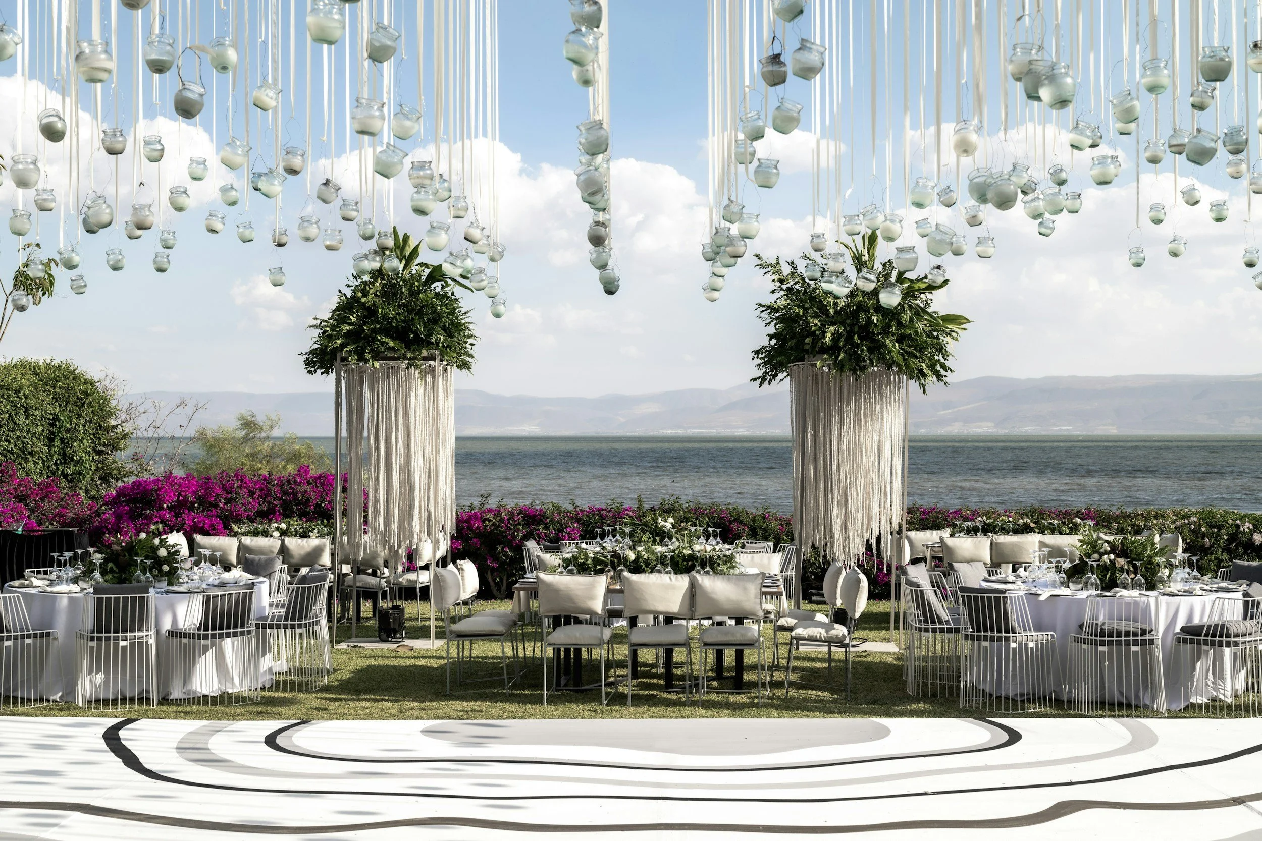 Beautiful outdoor wedding setup with a lakeside view, decorated with white chairs, large floral arrangements, and hanging glass jar decorations. The scene features a clear blue sky with clouds.