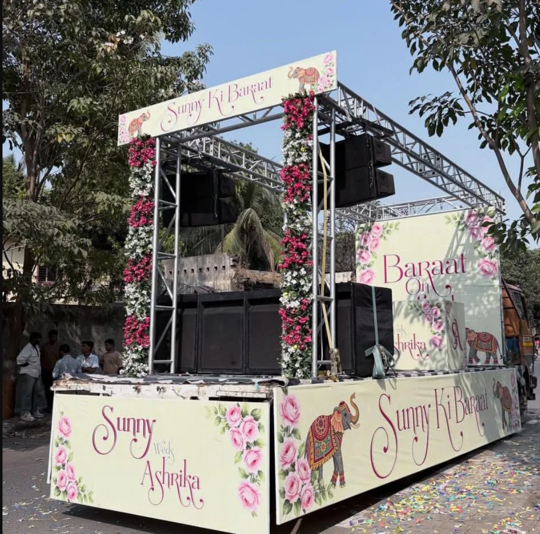 A decorated float for a wedding celebration with signs reading "Sunny Ki Baraat" and "Baraat on Ashrika." The float is adorned with pink and white flowers and elephant illustrations, and there are people in the background.