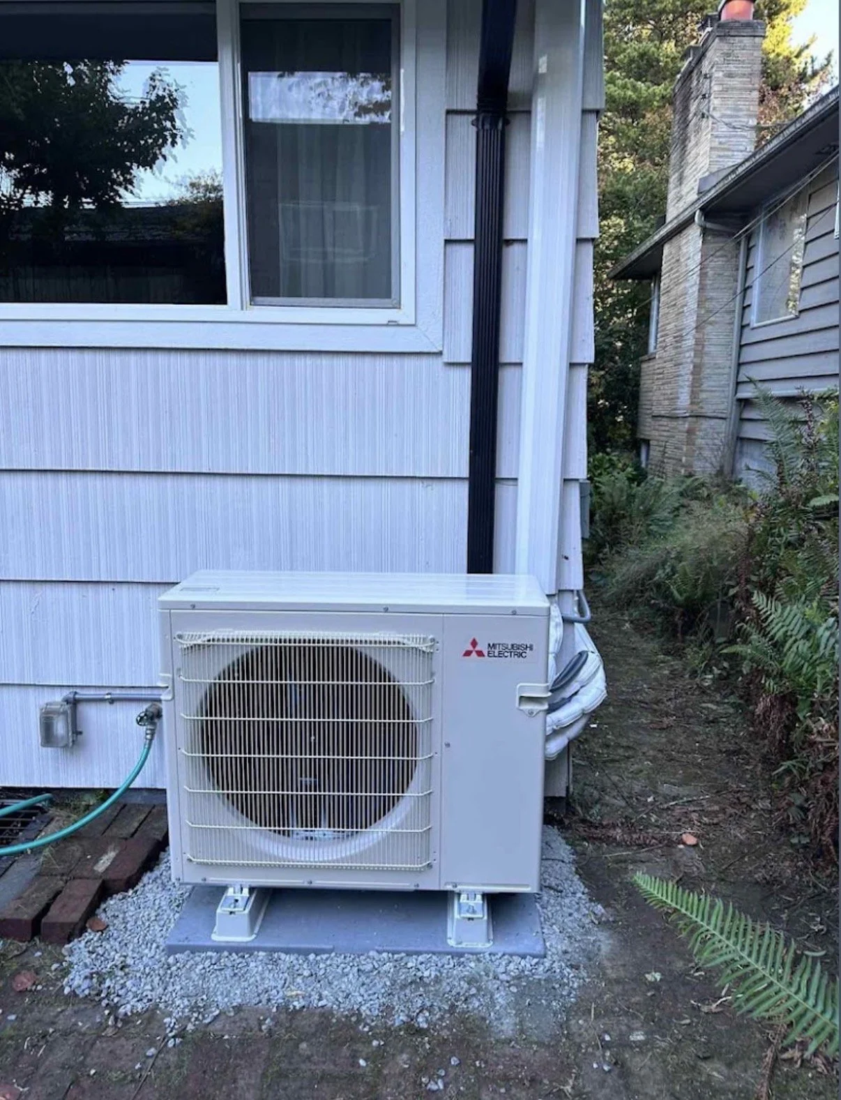 Outside view of an air conditioning or heat pump unit installed near the side of a house with light blue siding, with a window above it, power and refrigerant lines connected, on a gravel pad, surrounded by yard and plants.