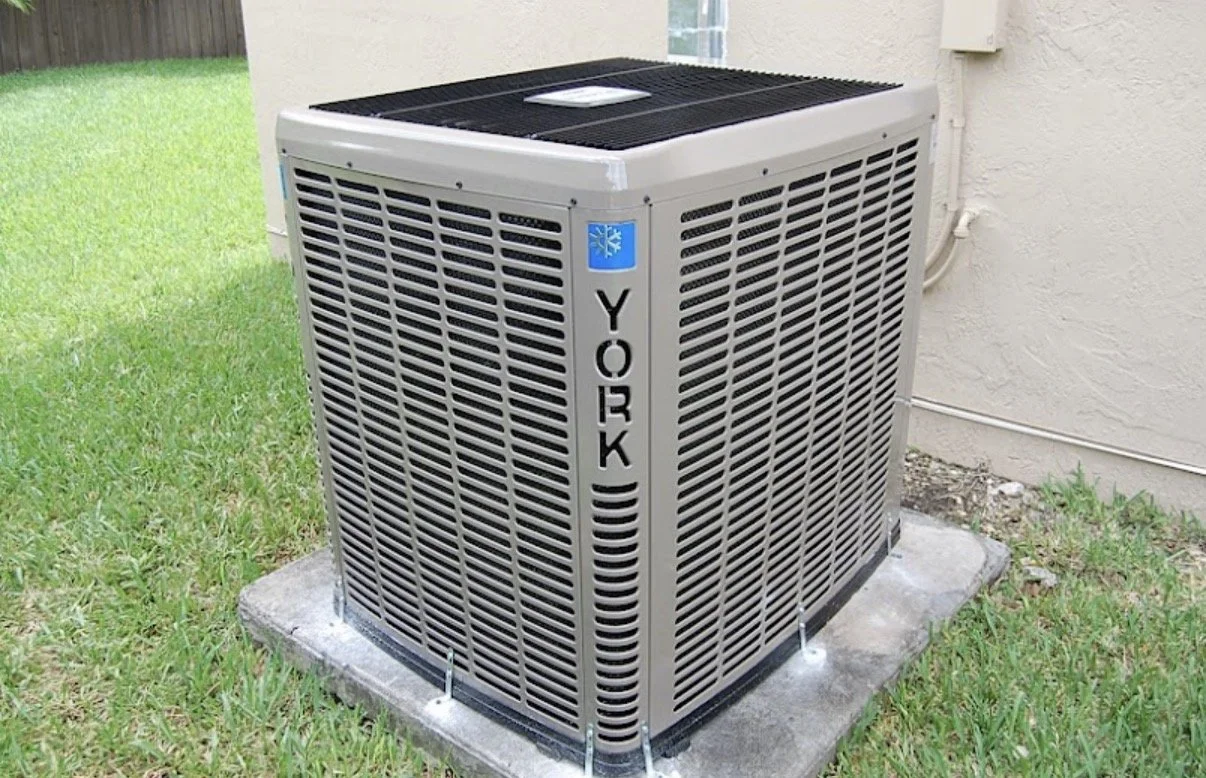 Outside view of a York air conditioning unit on a concrete slab next to a house with a beige wall and grass.