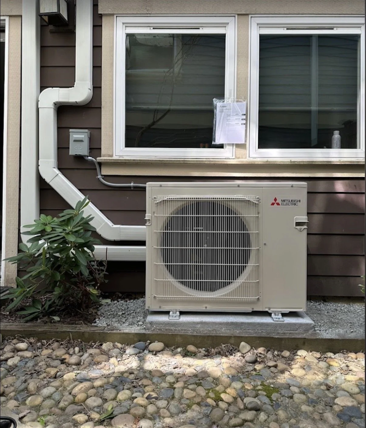 Exterior of a house featuring a Mitsubishi Electric air conditioning unit outside the window, with pipes and a small plant nearby.