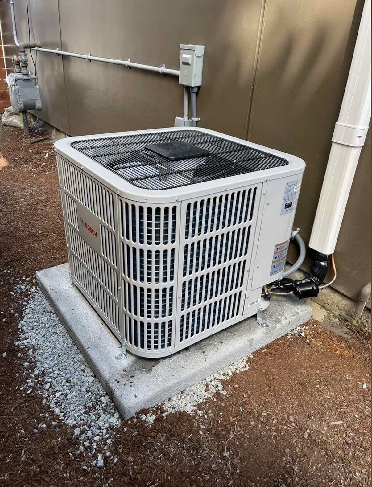 An outdoor Bosch air conditioning unit installed on a concrete slab with wiring and pipes connected to a building wall.
