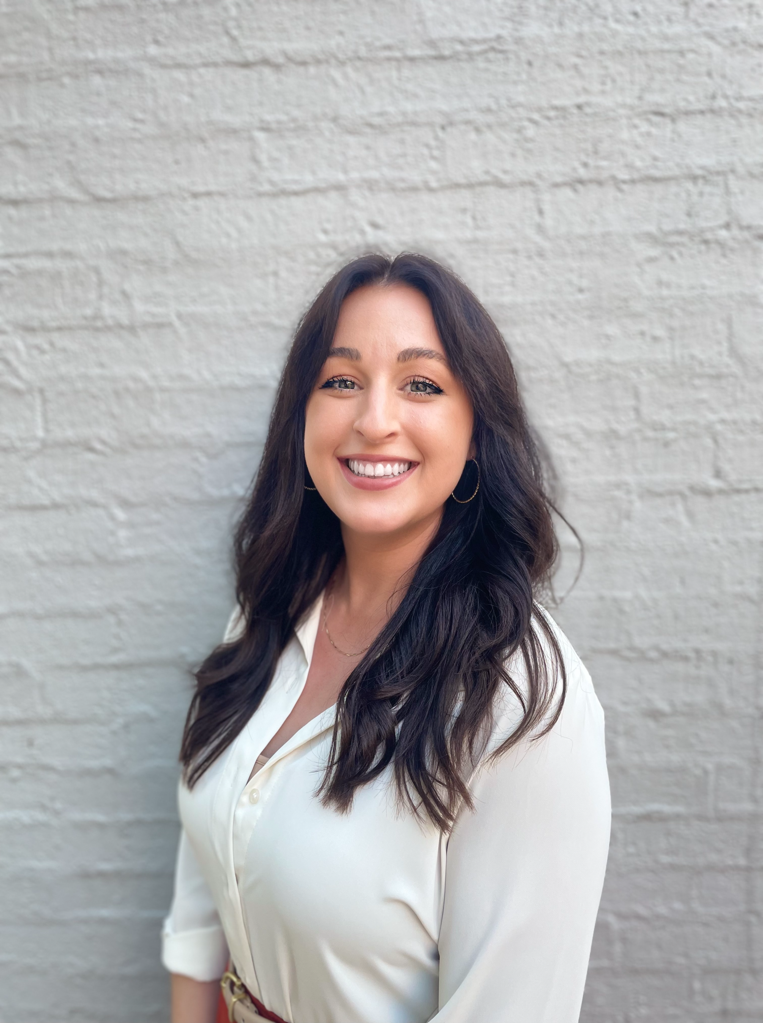 a young professional woman with long dark hair and white shirt smiling against a white brick building