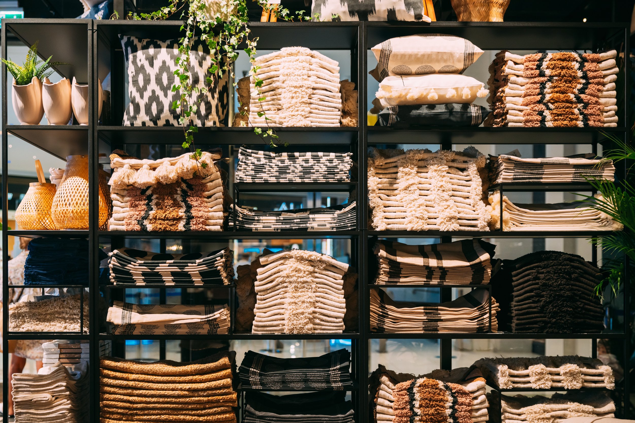 A black shelving unit holding neatly folded towels, pillowcases, and decorative items such as vases and plants, with a shopping mall in the background.