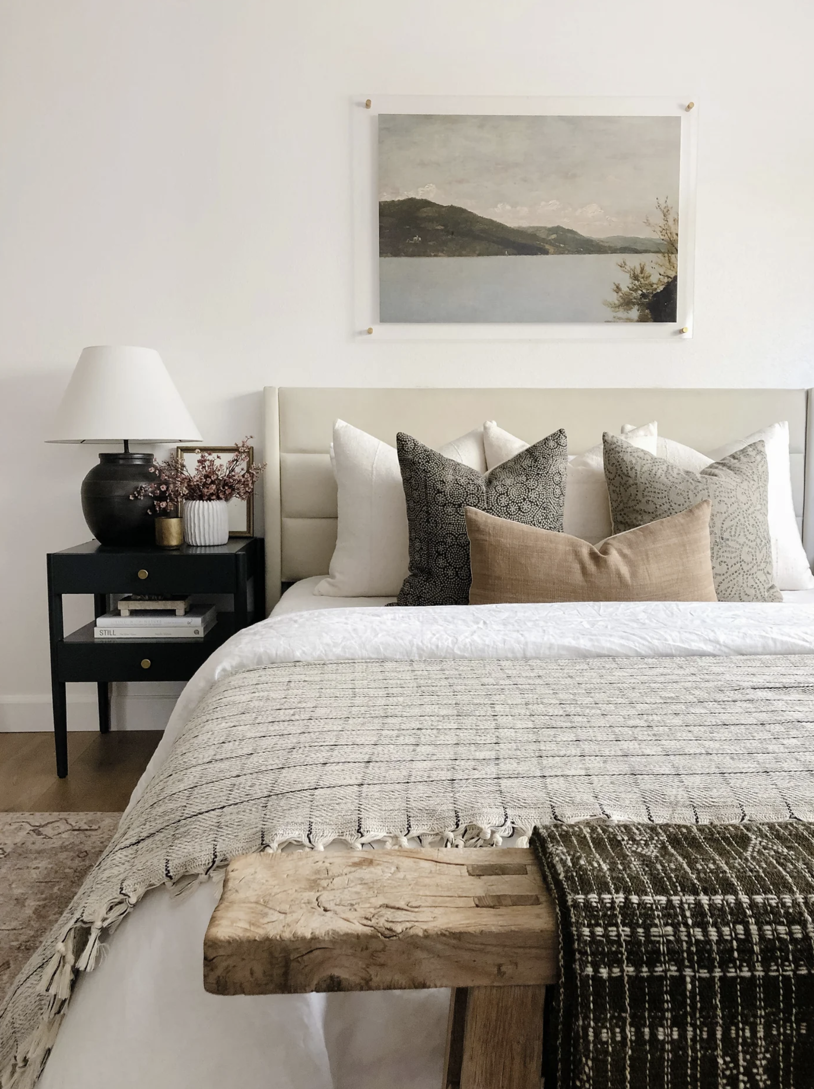 A bedroom with a variety of pillows on a bed, a black nightstand with decorative items, a lamp, a large landscape painting on the wall, and a wooden bench at the foot of the bed.