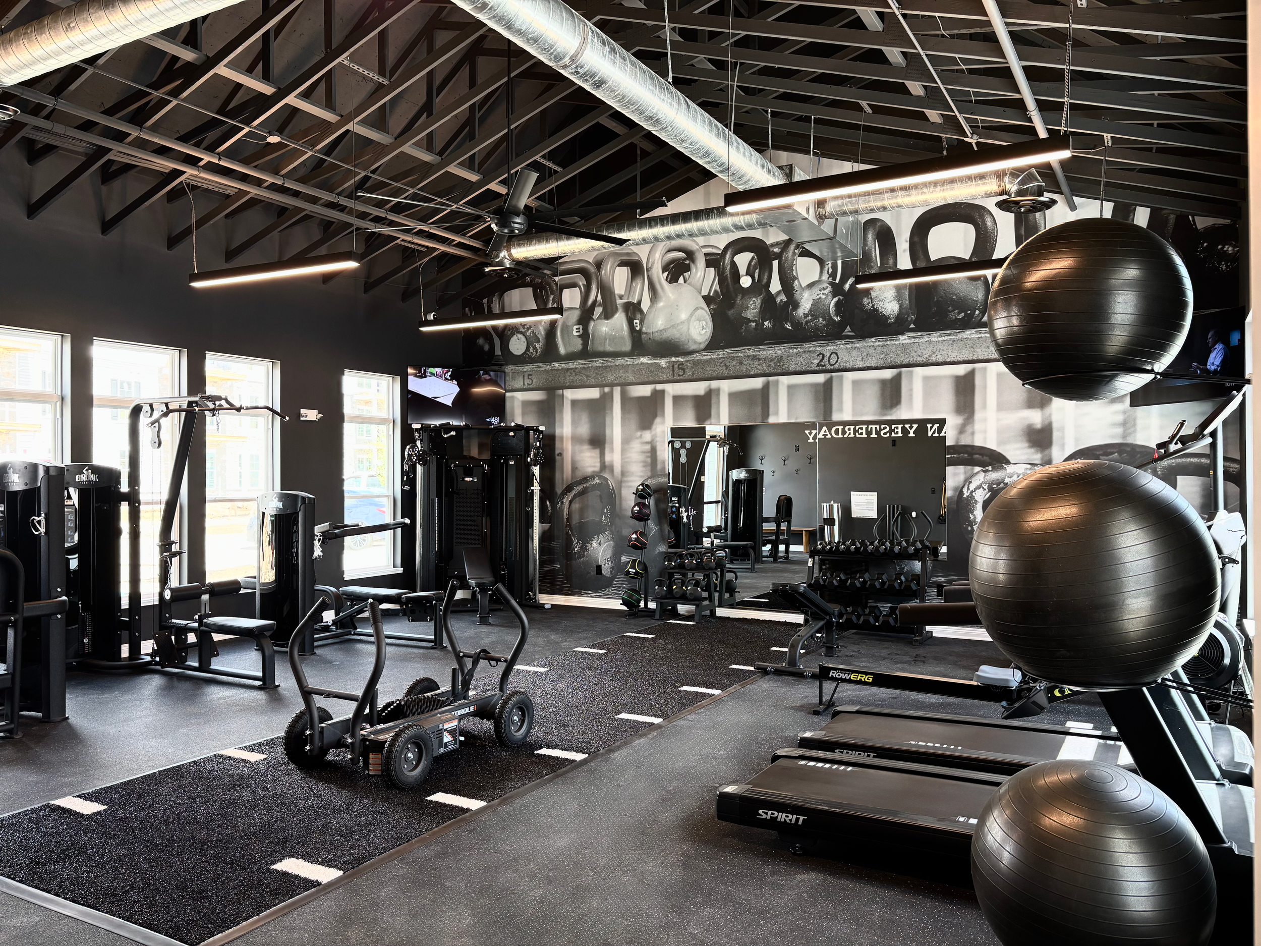 Interior of a fitness gym with various equipment including kettlebells, stability balls, treadmills, and weight machines. Large kettlebells are lined up on a shelf, and a large black exercise ball is hanging from the ceiling. The gym has black walls,