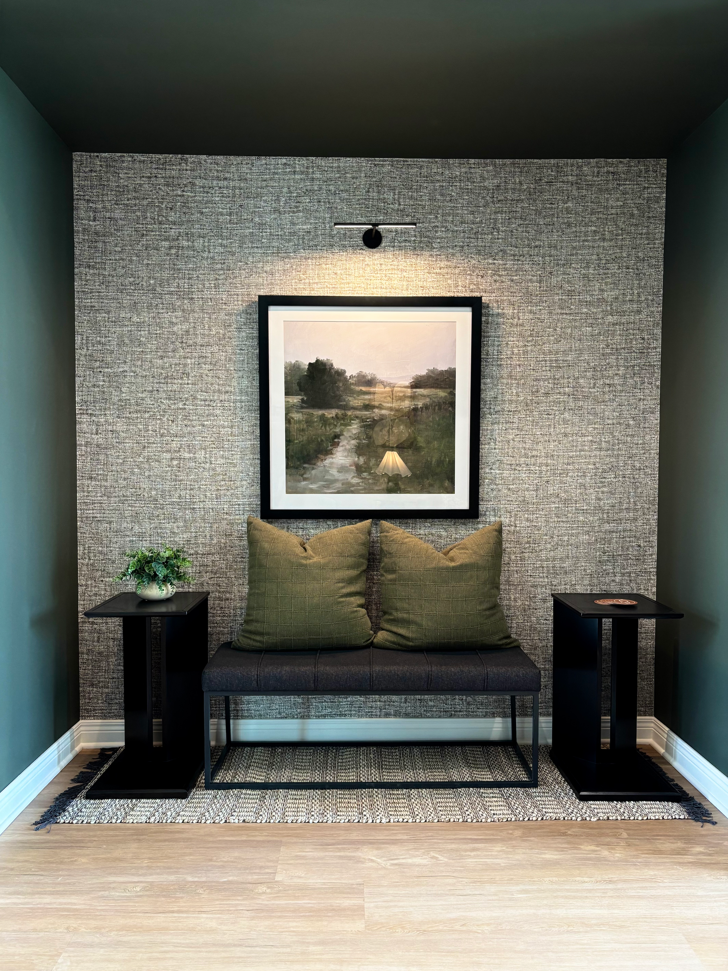 A modern interior space featuring a dark bench with two green pillows, a textured gray accent wall, and two black side tables with a small plant and decorative item, framed artwork, and carpet on a light wood floor.