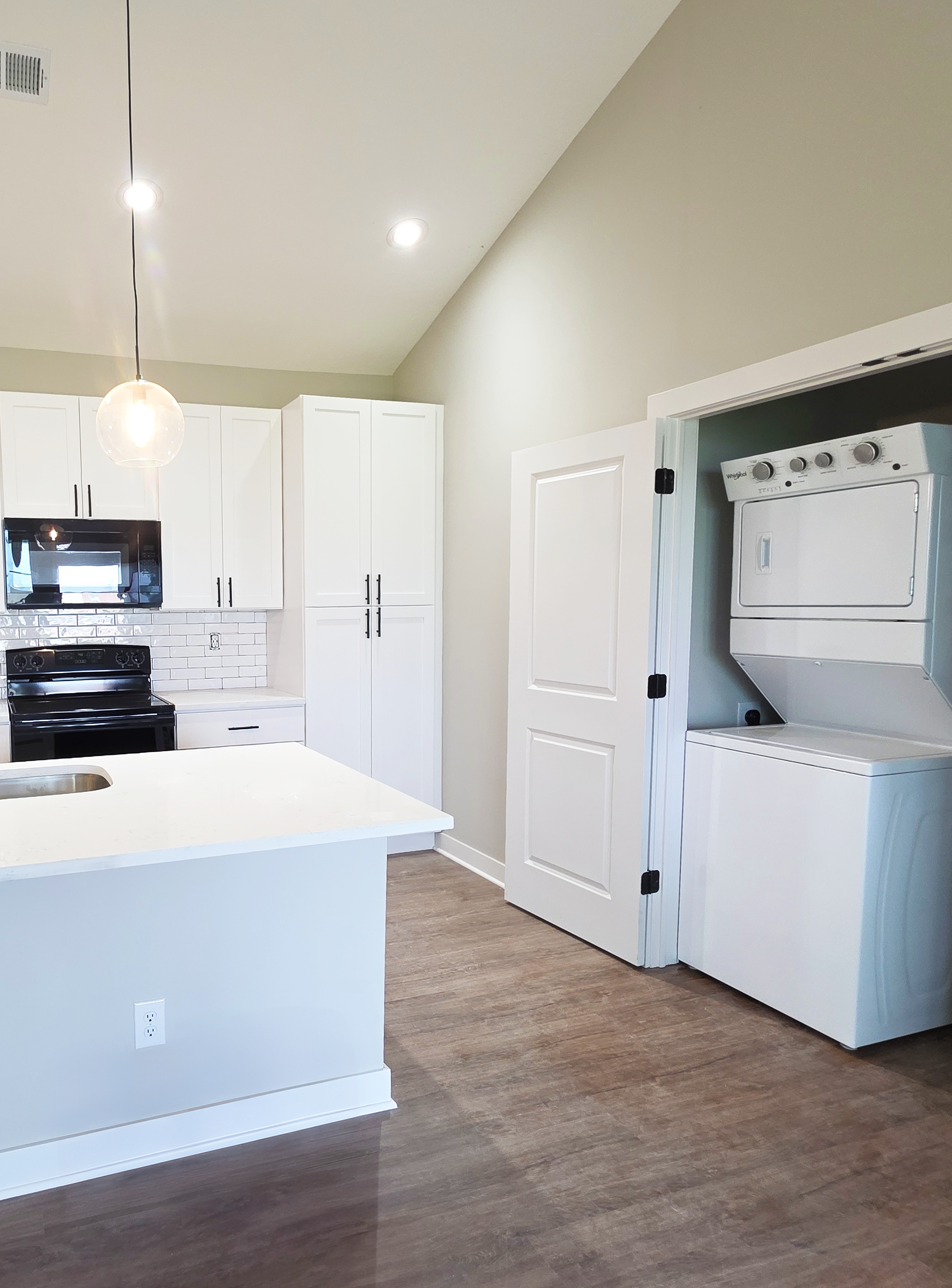Modern kitchen with white cabinets, a black stove, a white countertop island, and a stacked white washer and dryer in a laundry closet. Wooden flooring and recessed ceiling lights.