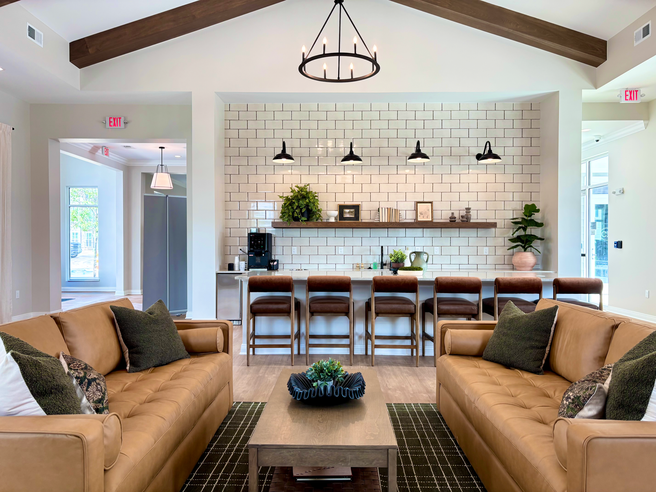 Living room with two tan sofas with dark cushion pillows, a wooden coffee table, and a black decorative tray with plants. In the background, there is a white brick wall with a wooden shelf holding plants, framed pictures, and decorative objects, and 
