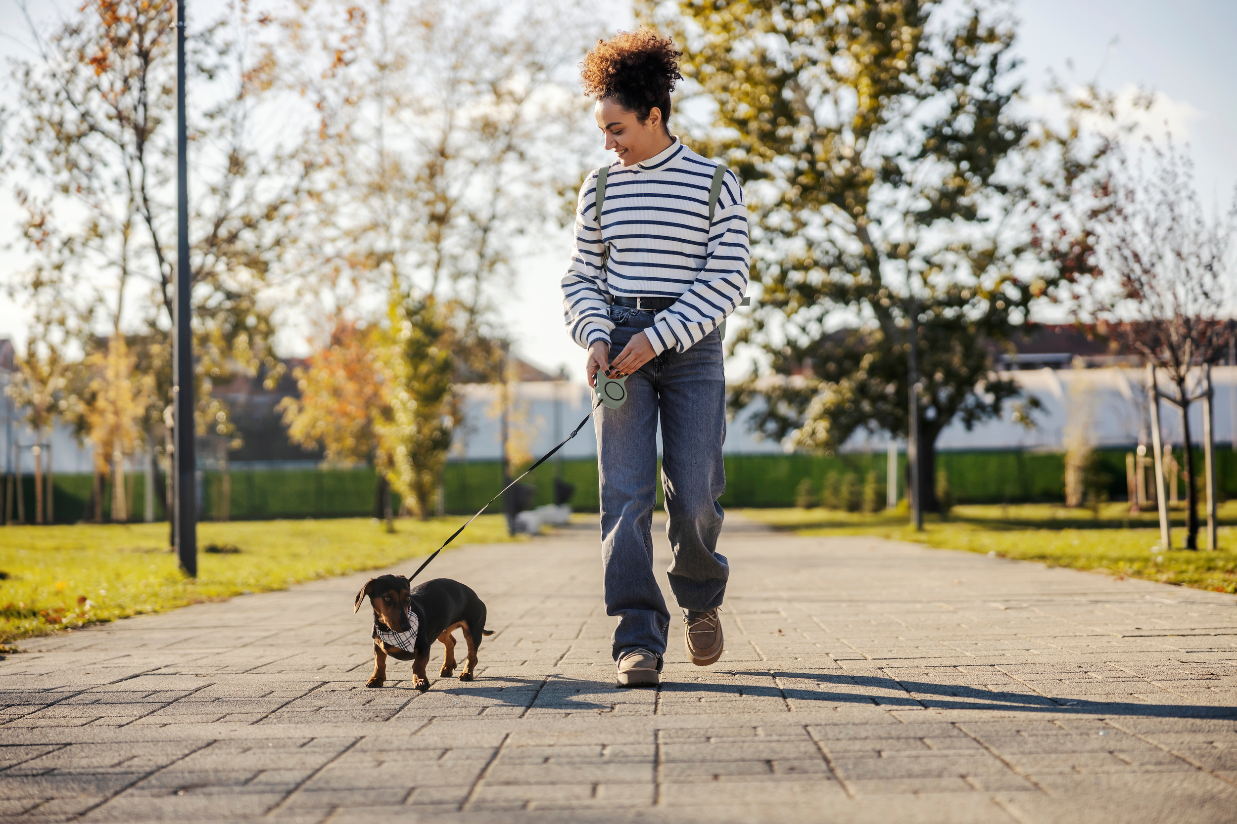 A young woman walking a small dog in a park during autumn. The woman is smiling, wearing a striped shirt and jeans, with a backpack, and the dog is on a leash wearing a bandana. The background has trees with fall leaves and a clear sky.