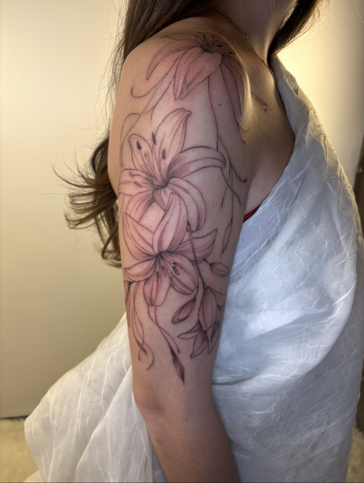 A person's upper arm and shoulder with an outline tattoo of lilies in progress.