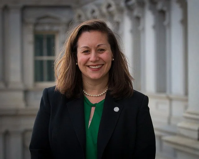 Fowler Federal, LLC founder, Dana Fowler. Image of a woman with shoulder-length brown hair smiling, wearing a dark blazer, green top, pearl necklace, standing outside a the Eisenhower Executive Office  Building with white columns.
