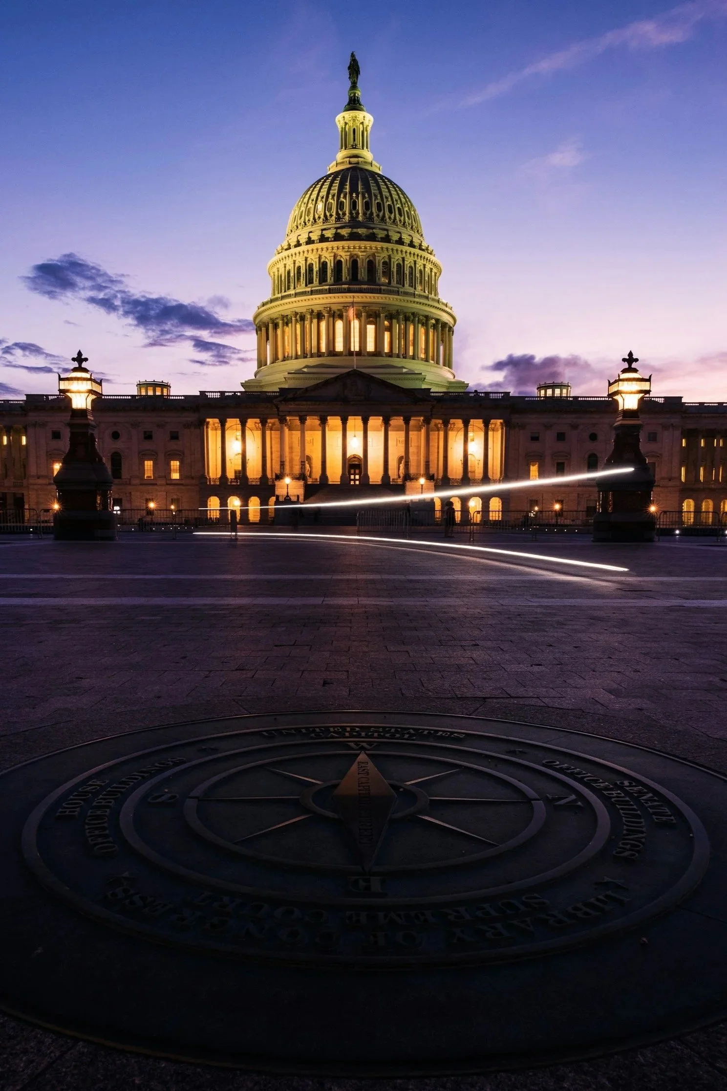 Nighttime view of the United States Capitol building with illuminated dome, set against a purple sky, with light trails from passing vehicles on the street in the foreground.
