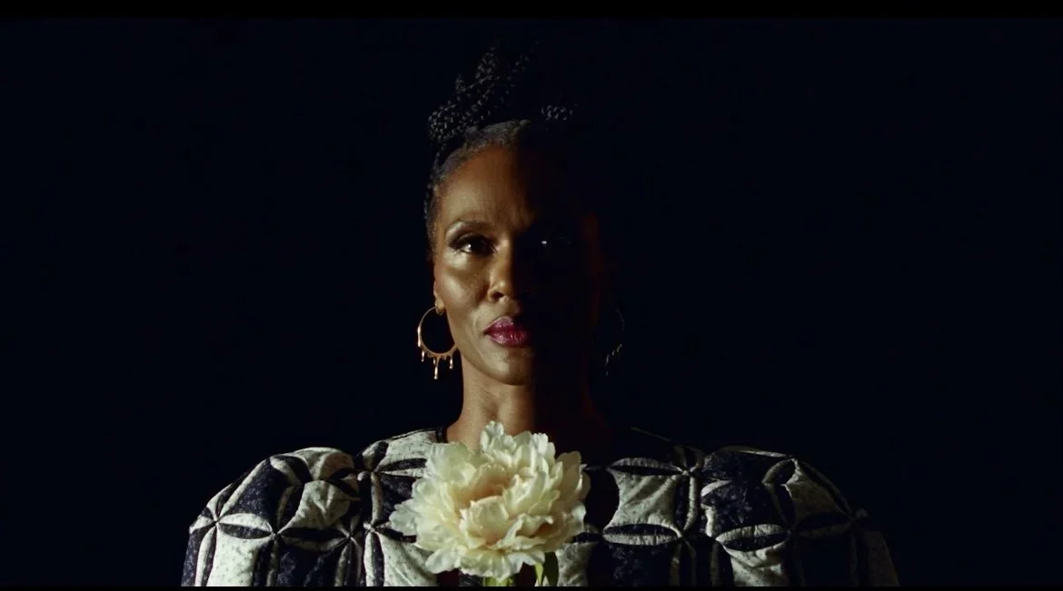 Portrait of a woman with dark skin, wearing a black and white patterned top, gold hoop earrings, holding a white flower, with lighting emphasizing half her face against a dark background.