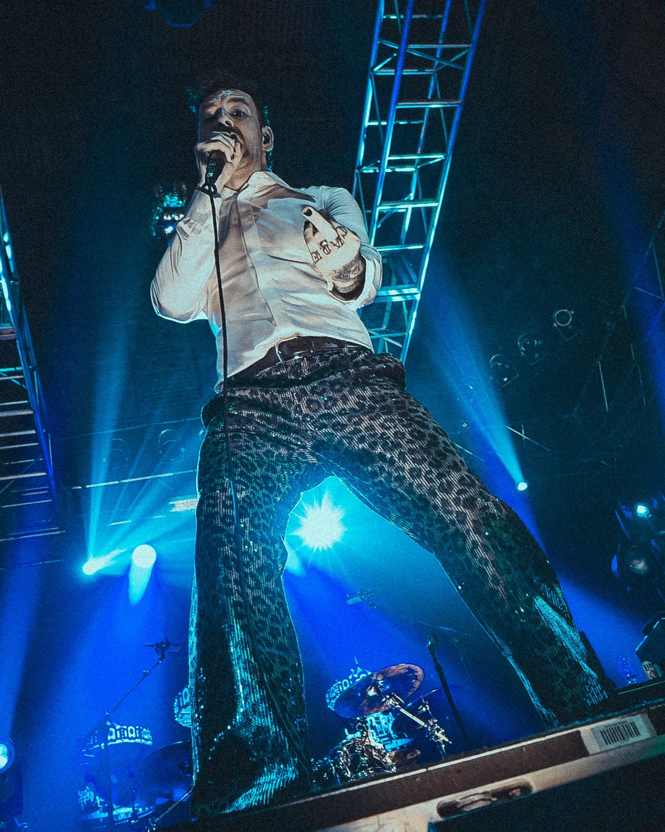 A male performer on stage holding a microphone, wearing a white shirt and leopard print pants, with stage lights and metal trusses in the background.