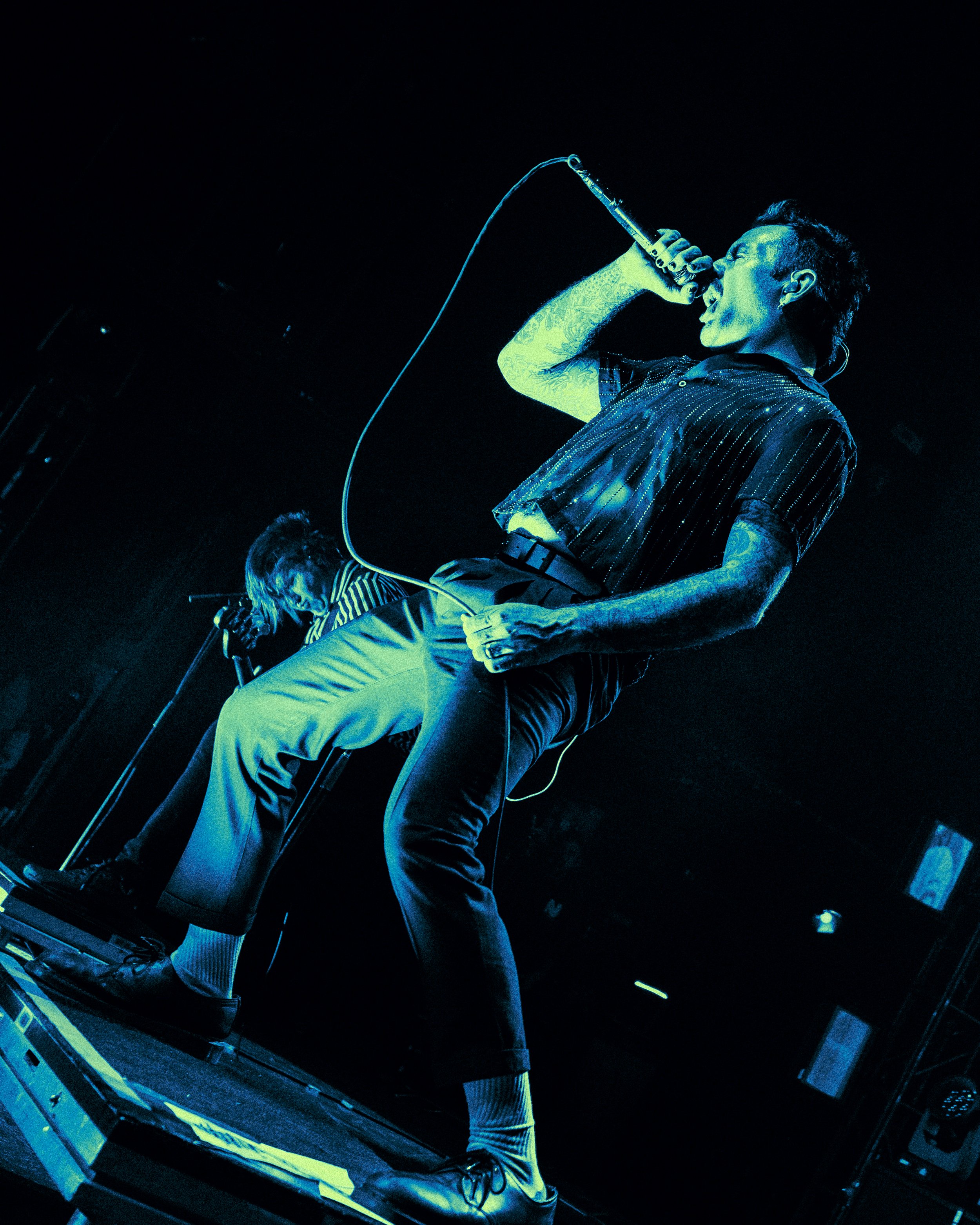 A male vocalist singing into a microphone on a dark stage, with a band member playing guitar in the background.