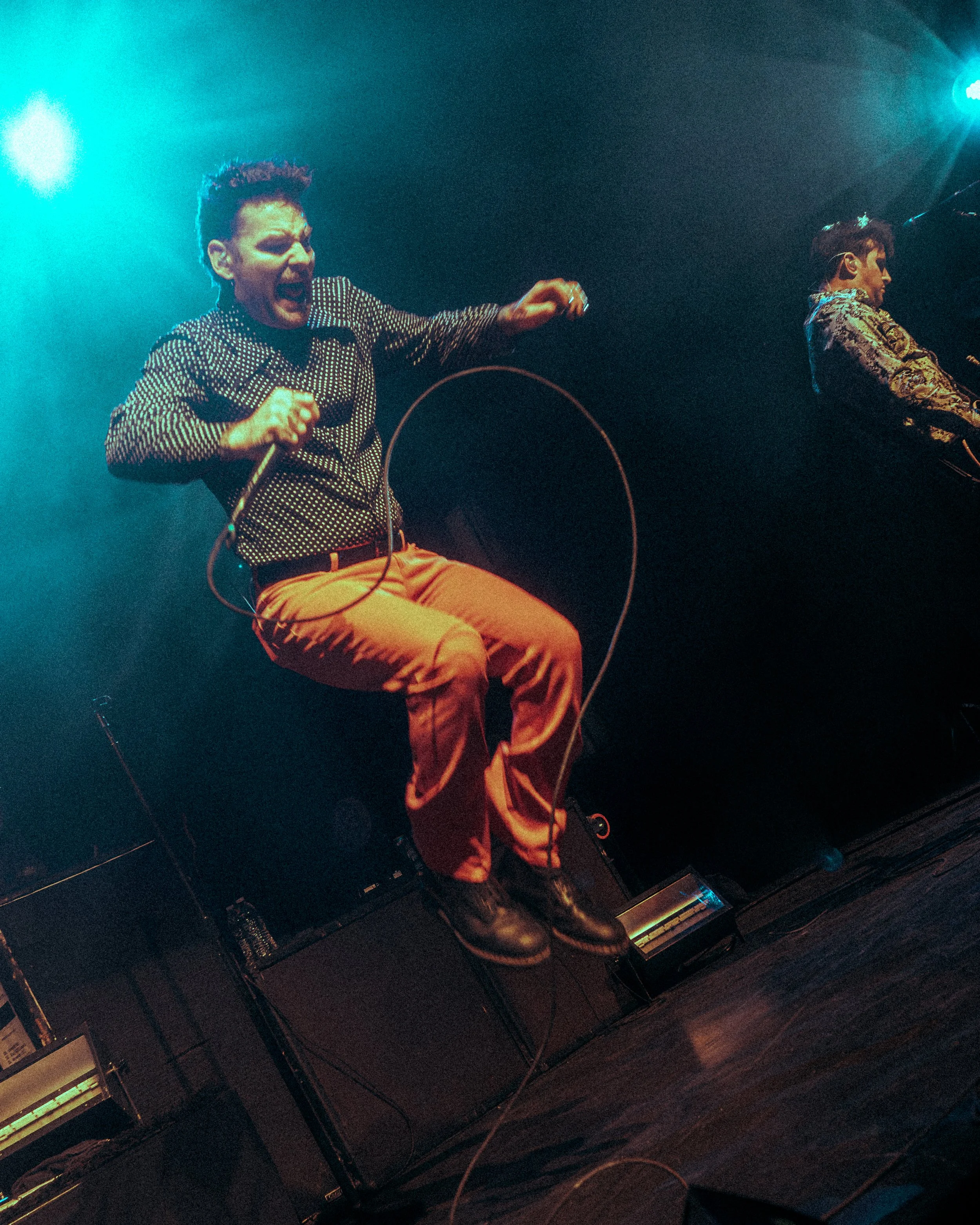 Two people are performing on stage with lighting creating an energetic atmosphere. The individual on the left has short hair, is wearing a polka-dot shirt and orange pants, and appears to be singing passionately into a microphone. The person on the r