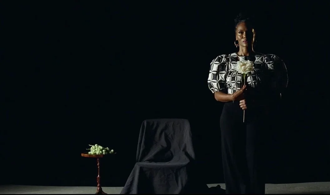 A woman standing against a black background holding a white flower. There is a chair covered with black fabric and a small table with grapes next to her.