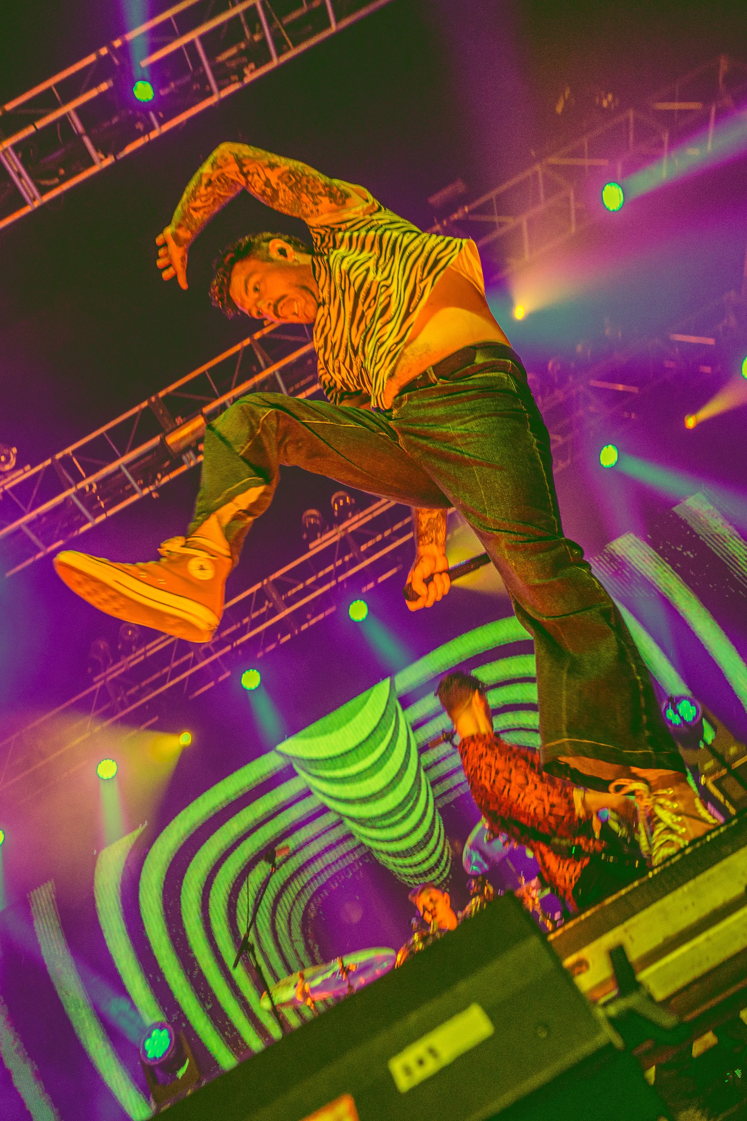 A performer on stage in a zebra-striped shirt and striped pants, jumping with a microphone in hand, under colorful stage lights with a curved, striped background and other dancers.