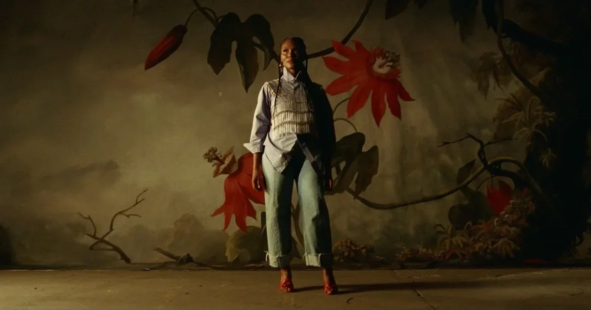 A woman standing in front of a mural with large red and orange flowers and green leaves, wearing a light-colored vest over a button-up shirt, cuffed jeans, and red shoes.