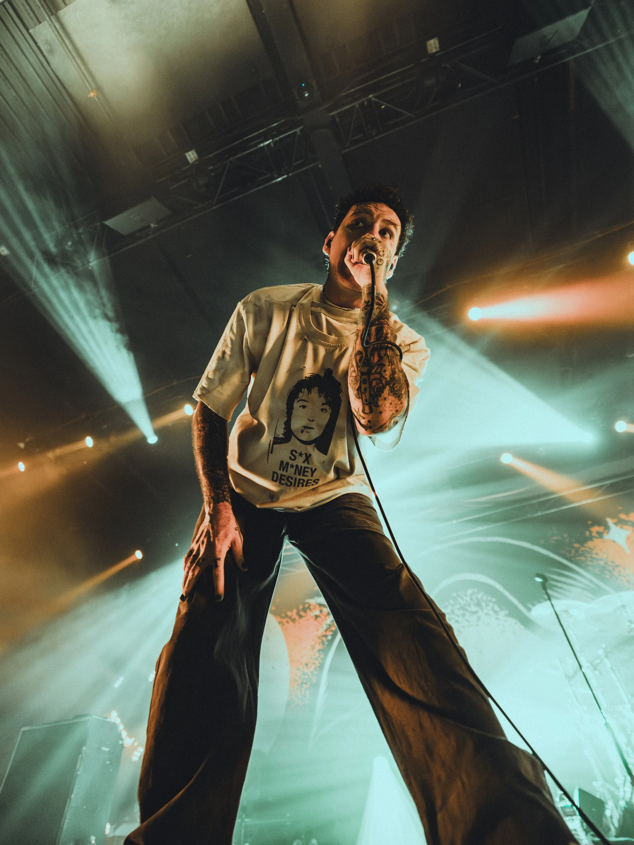 A male performer with tattoos wearing a graphic T-shirt, holding a microphone, on stage with colorful stage lighting and a dark background.