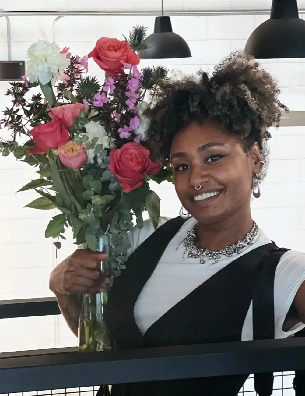 A woman with curly hair and jewelry holding a large bouquet of colorful flowers, smiling indoors.