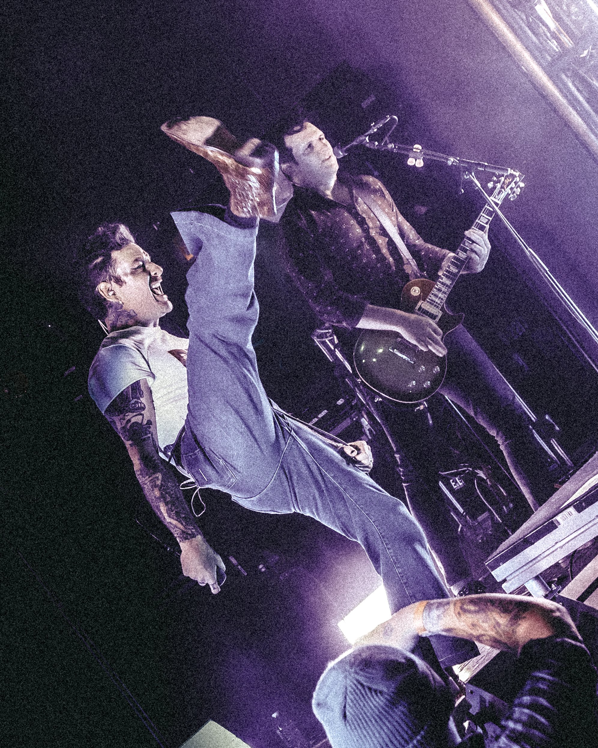 Two musicians perform on stage, one playing a guitar and singing into a microphone, the other with tattoos singing passionately.