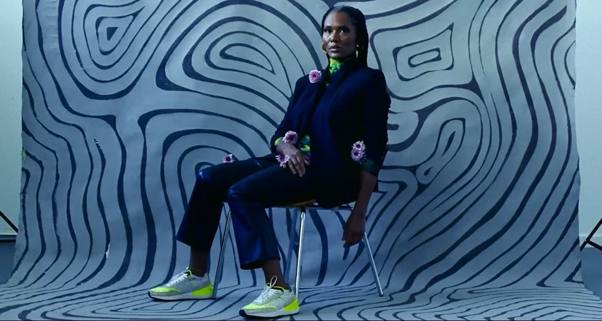 A woman sitting on a chair in front of a blue and black abstract patterned backdrop, wearing a dark blazer, black pants, and bright yellow and white sneakers, with flowers as accessories.
