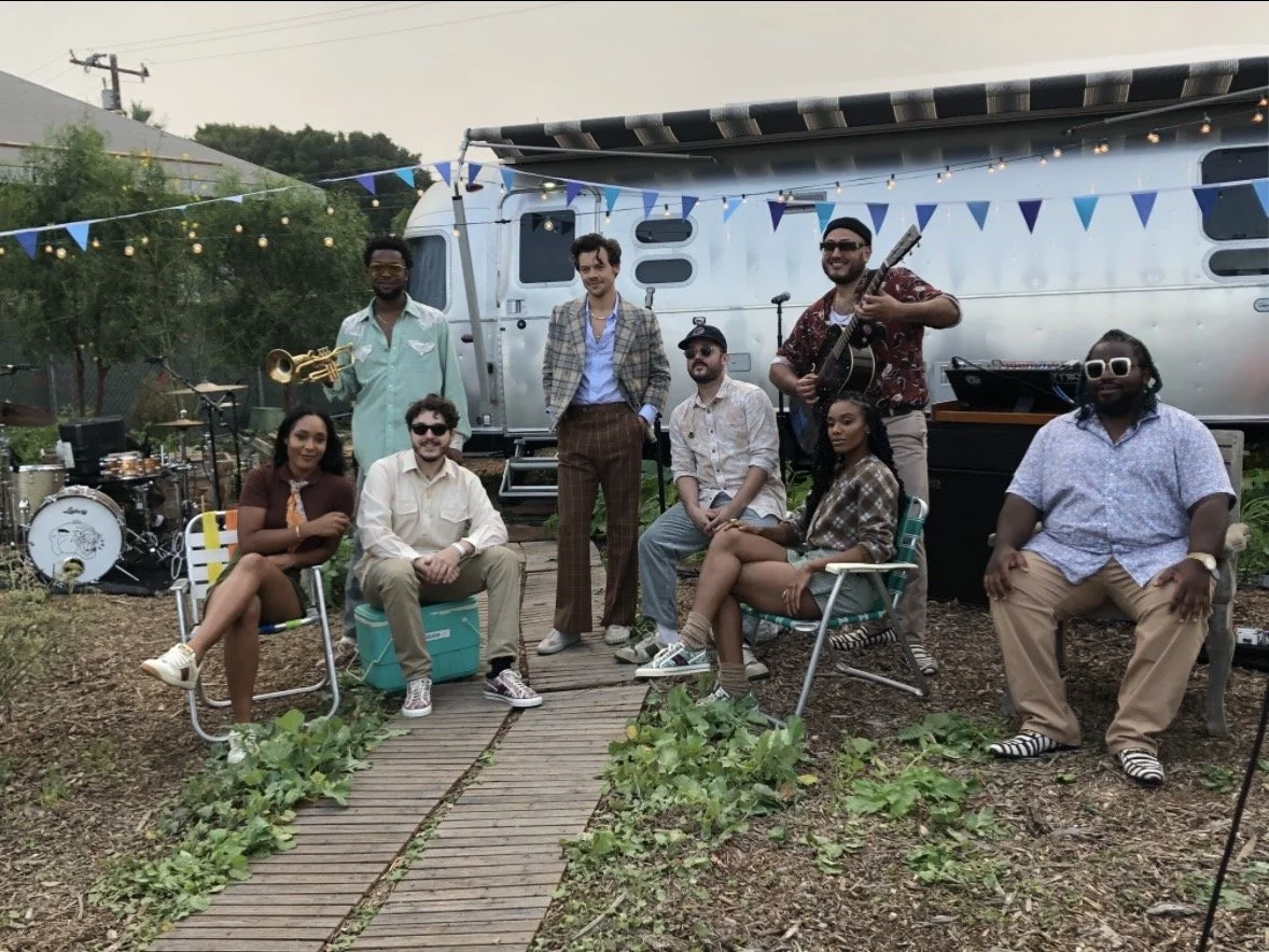A group of nine people gathered outdoors at a casual event, with musical instruments like drums, a saxophone, and a guitar, standing and sitting on a wooden pathway surrounded by greenery, with a trailer decorated with string lights and blue banners 