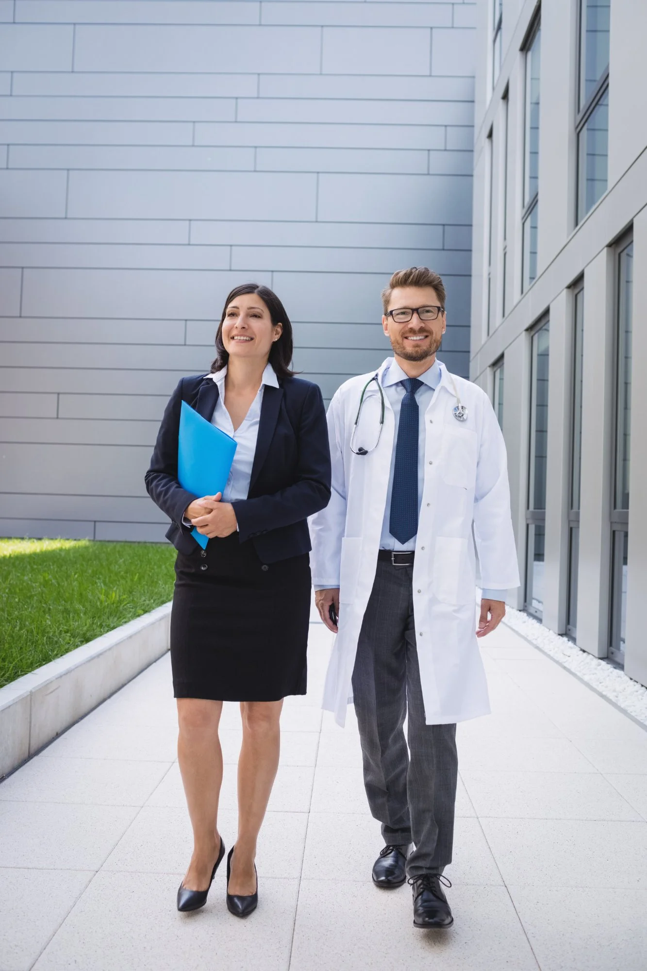 A businessman and a female doctor walking outside a modern building, smiling, with the businessman wearing glasses and a white coat.