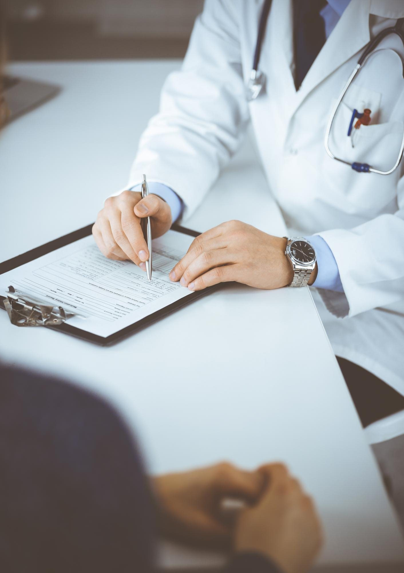 A person in a white lab coat and stethoscope appears to be a medical professional filling out a form or document on a clipboard on a white table.