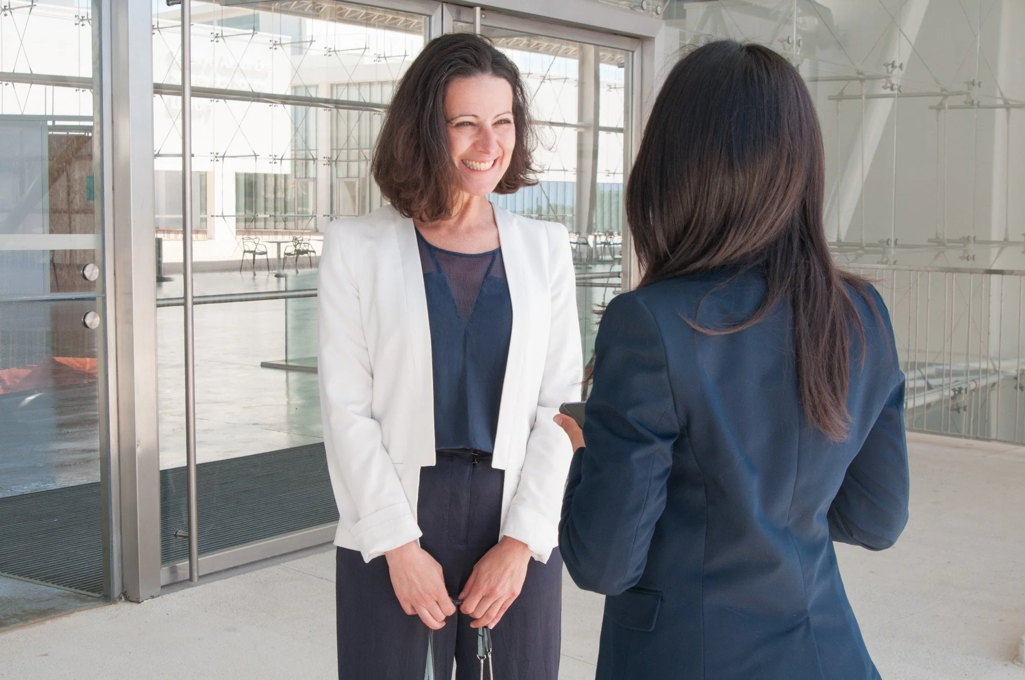 Two women engaged in conversation near the entrance of a modern building, with large glass windows and doors.